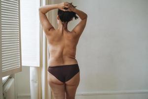 selbstliebende attraktive Frau plus Größe in Unterwäsche von hinten in der Nähe des Fensters im hellen Raum, Körperliebe foto