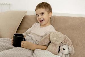 entzückender kleiner junge, der zu hause heißen tee auf dem bett trinkt und sich entspannt. Kind ruht, in eine Decke gehüllt, mit Becher in seinem Zimmer. Interieur und Kleidung in natürlichen Erdfarben. gemütliche Umgebung. foto