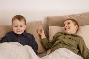 zwei kleine und süße kaukasische jungen, die zu hause zusammen auf dem bett spielen. Interieur und Kleidung in natürlichen Erdfarben. gemütliche Umgebung. Kinder haben Spaß, zwei Brüder. foto