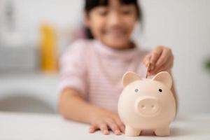 kleines asiatisches mädchen, das geld in einem sparschwein spart, über sparen lernt, kind spart geld für zukünftige bildung. geld, finanzen, versicherungen und personenkonzept foto