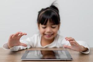 Asiatisches kleines süßes Mädchen, das den digitalen Tablet-Bildschirm auf dem Tisch berührt foto
