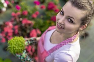 junge Frau in einer Gärtnerei, die eine Blumenpflanze in ihren Händen hält, während sie auf dem Gehweg zwischen Pflanzen kniet. foto