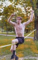 junger Mann balanciert und springt auf Slackline. Mann geht, springt und balanciert auf einem Seil im Park. foto