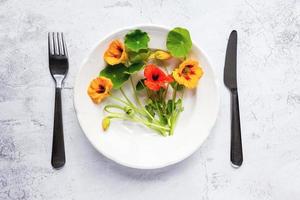 Kapuzinerkresse-Pflanzen, indische Kresseblüten und -blätter in Teller, Besteck auf weißem Tisch, Overhead-Flachlage foto