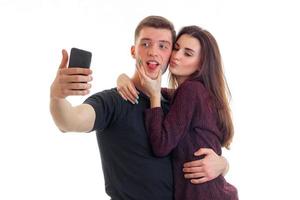 Schönes Mädchen küsst einen jungen attraktiven Mann auf die Wange und er macht Selfi am Telefon foto