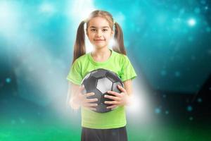 und lächelt auf Kamerakleines Mädchen im grünen Hemd, das einen Fußball hält foto