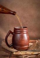 Bierlehm brauner Krug mit Bier auf einem Holztisch auf dunklem Hintergrund. ein strahl helles bier ergießt sich in ein glas foto