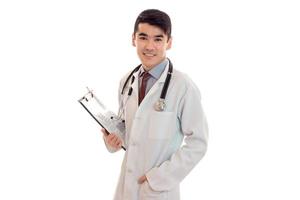 junge elegante brünette Ärztin in Uniform mit Stethoskop lächelt in die Kamera und macht sich Notizen isoliert auf weißem Hintergrund foto