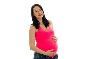 Porträt der jungen schönen schwangeren Frau in rosa Hemd, die ihren Bauch berührt und vor der Kamera lächelt, isoliert auf weißem Hintergrund foto