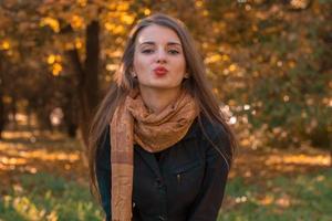 schönes Mädchen steht im Herbstpark in einem langen Schal, der direkt schaut und Kuss sendet foto