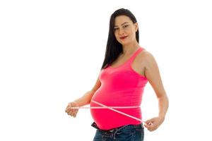 schöne junge schwangere brünette Frau, die ihren Bauch berührt, posiert mit Maßband und schaut isoliert auf weißem Hintergrund in die Kamera foto