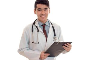 Porträt eines jungen, fröhlichen, brünetten Arztes in weißer Uniform, der mit einem Stethoskop am Hals vor der Kamera lächelt, macht Notizen, die auf leerem Hintergrund isoliert sind foto