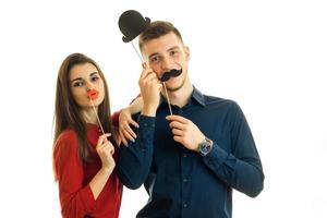 Junges schönes Mädchen und Kerl halten in der Nähe von Gesichtspapierstützen in Form von Schnurrbart, Lippen und Hut und posieren für die Kamera foto