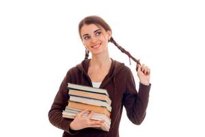Fröhliches, intelligentes Studentenmädchen in brauner Sportkleidung mit vielen Büchern in der einen Hand, die ihr Haar berührt und isoliert auf weißem Hintergrund lächelt foto