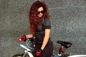 Glamour-Frau mit Sonnenbrille mit lockigem rotem Haar auf dem Fahrrad foto