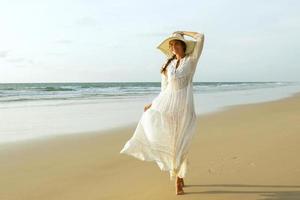 frau, die schönes weißes kleid trägt, geht während des sonnenuntergangs am strand spazieren foto