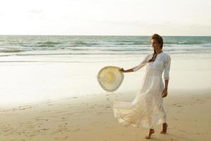 frau, die schönes weißes kleid trägt, geht während des sonnenuntergangs am strand spazieren foto