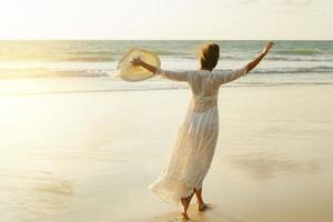 frau, die schönes weißes kleid trägt, geht während des sonnenuntergangs am strand spazieren foto