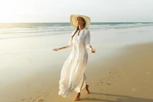 frau, die schönes weißes kleid trägt, geht während des sonnenuntergangs am strand spazieren foto