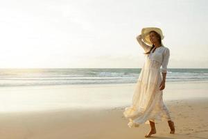 frau, die schönes weißes kleid trägt, geht während des sonnenuntergangs am strand spazieren foto