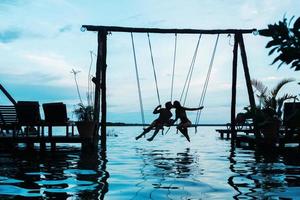 Silhouette eines Paares auf Schaukeln, das über dem Wasser liegt foto