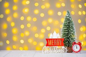 weihnachtsbaum, rote uhr und holzbeschriftung frohe weihnachten auf hellem bokeh hintergrund. foto