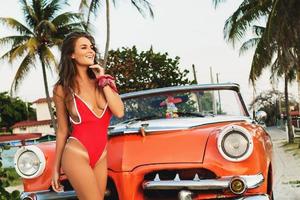 sexy Frau mit rotem Badeanzug posiert neben einem Retro-Cabriolet-Auto am Strand foto
