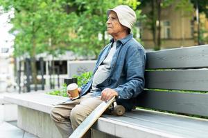 Älterer Mann mit Longboard sitzt auf der Bank und trinkt Kaffee in einem Stadtpark foto
