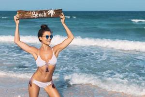 sexy Frau trägt Badeanzug mit altem Holzschild am Strand foto