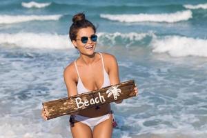 sexy Frau trägt Badeanzug mit altem Holzschild am Strand foto