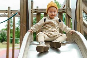 Süßer kleiner Junge, der auf einer Spielplatzrutsche im öffentlichen Park sitzt foto