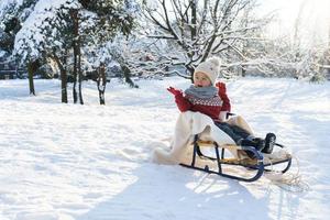 Kleinkindjunge, der an sonnigen Wintertagen auf dem Schlitten in einem verschneiten Stadtpark sitzt foto