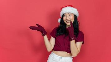 Foto positiv Mädchen in Weihnachtsmann-Hut halten Hand demonstrieren Weihnachten Neujahr Anzeige Promo Touch Finger Zähne tragen warme Wintersaison Kleidung isoliert roter Hintergrund