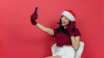 Foto einer charmanten Dame, die ein Telefon hält und Selfies macht, die über Skype winken und mit der Handfläche Hallo sagen, Sitzkomfort auf rotem Hintergrund. konzept des neuen jahres und des weihnachtstages.