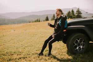 junger Mann entspannt und trinkt Kaffee an der Motorhaube des Geländefahrzeugs auf dem Land foto