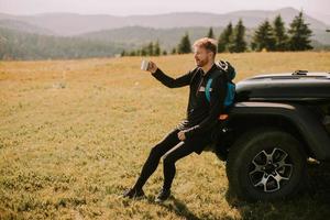 junger Mann entspannt und trinkt Kaffee an der Motorhaube des Geländefahrzeugs auf dem Land foto