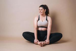 Selbstliebende junge Frau plus Größe in Sportbekleidung beim Stretching auf beigem Hintergrund, Körperliebe foto