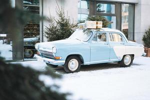 Hellblaues Oldtimerauto mit natürlichem Weihnachtsbaum und Kisten mit Geschenken auf der Winterstraße foto