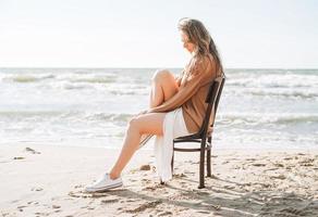 junge sorglose schöne Frau mit langen Haaren im Pullover, die auf einem Stuhl am Meeresstrand sitzt foto