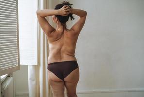 selbstliebende attraktive Frau plus Größe in Unterwäsche von hinten in der Nähe des Fensters im hellen Raum, Körperliebe foto