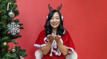 hübsche lächelnde asiatische frau in sankt-kostüm und weihnachtsmütze wünscht sich neben weihnachtsbaum auf rotem hintergrund für die saisonfeier foto