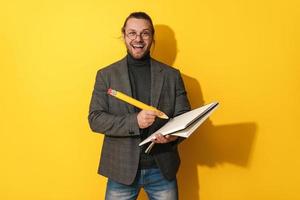 Fröhlicher Mann mit Brille, der großen Bleistift und Notizbuch auf gelbem Hintergrund hält foto