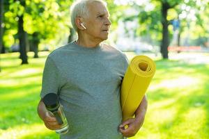 aktiver älterer mann mit einer übungsmatte und einer flasche wasser im grünen stadtpark foto