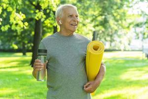 aktiver älterer mann mit einer übungsmatte und einer flasche wasser im grünen stadtpark foto