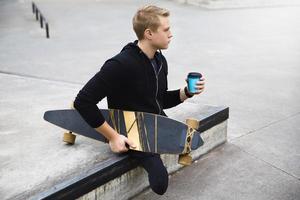 Behinderter Typ mit einer Tasse Kaffee vor dem Longboard-Fahren in einem Skatepark foto