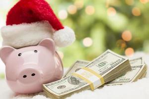Rosa Sparschwein mit Weihnachtsmütze in der Nähe von Geldstapeln auf Schneeflocken foto