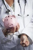 Arzt in Handschellen hält Sparschwein mit Laborkittel, Stethoskop foto