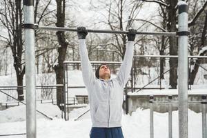 sportlicher Mann, der während seines Wintertrainings im Freien Klimmzüge an der horizontalen Stange macht foto