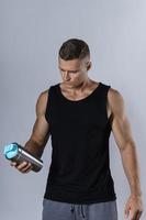 Mann, der ein leeres schwarzes Tanktop trägt, trinkt Protein oder andere Ergänzungen aus einem Metallshaker foto