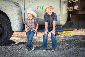 Zwei Jungen mit Cowboyhüten, die sich in einer rustikalen ländlichen Umgebung an einen antiken Lastwagen lehnen. foto